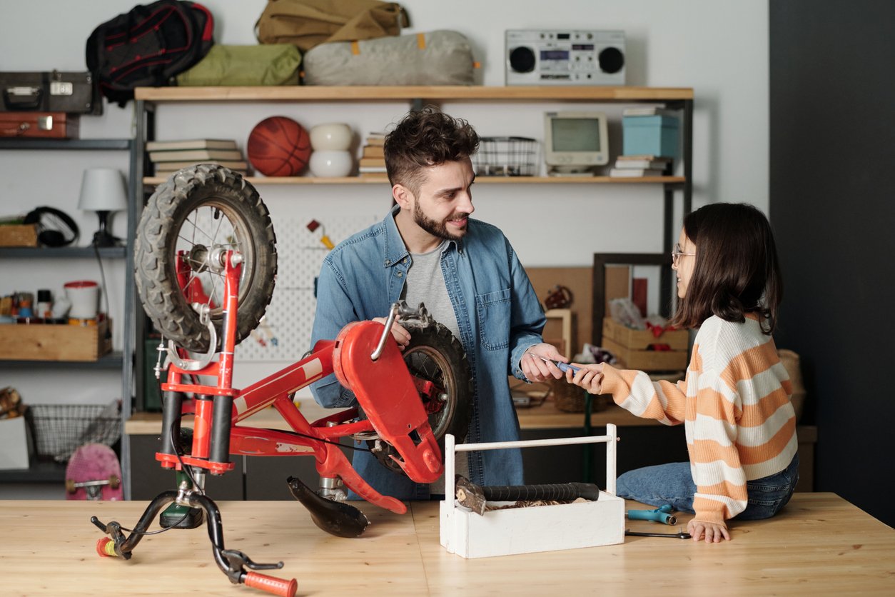 Young man taking handtool held by his daughter while repairing her bicycle