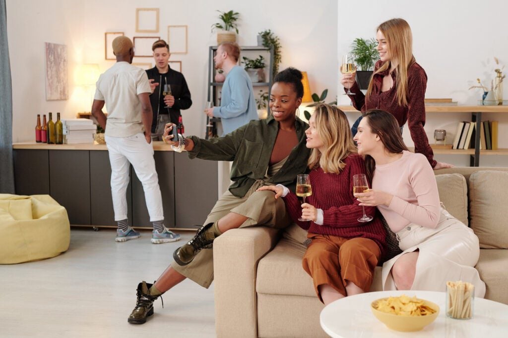 Happy young intercultural women with flutes of champagne looking at you with toothy smiles while enjoying home party against their boyfriends